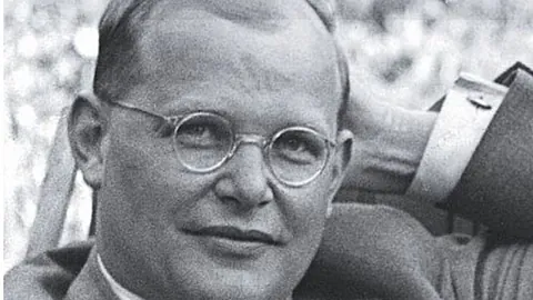 Schwarz-weiß-Fotografie von Dietrich Bonhoeffer, der mit Brille und Anzug entspannt in die Kamera blickt. Eine Hand liegt lässig hinter seinem Kopf.